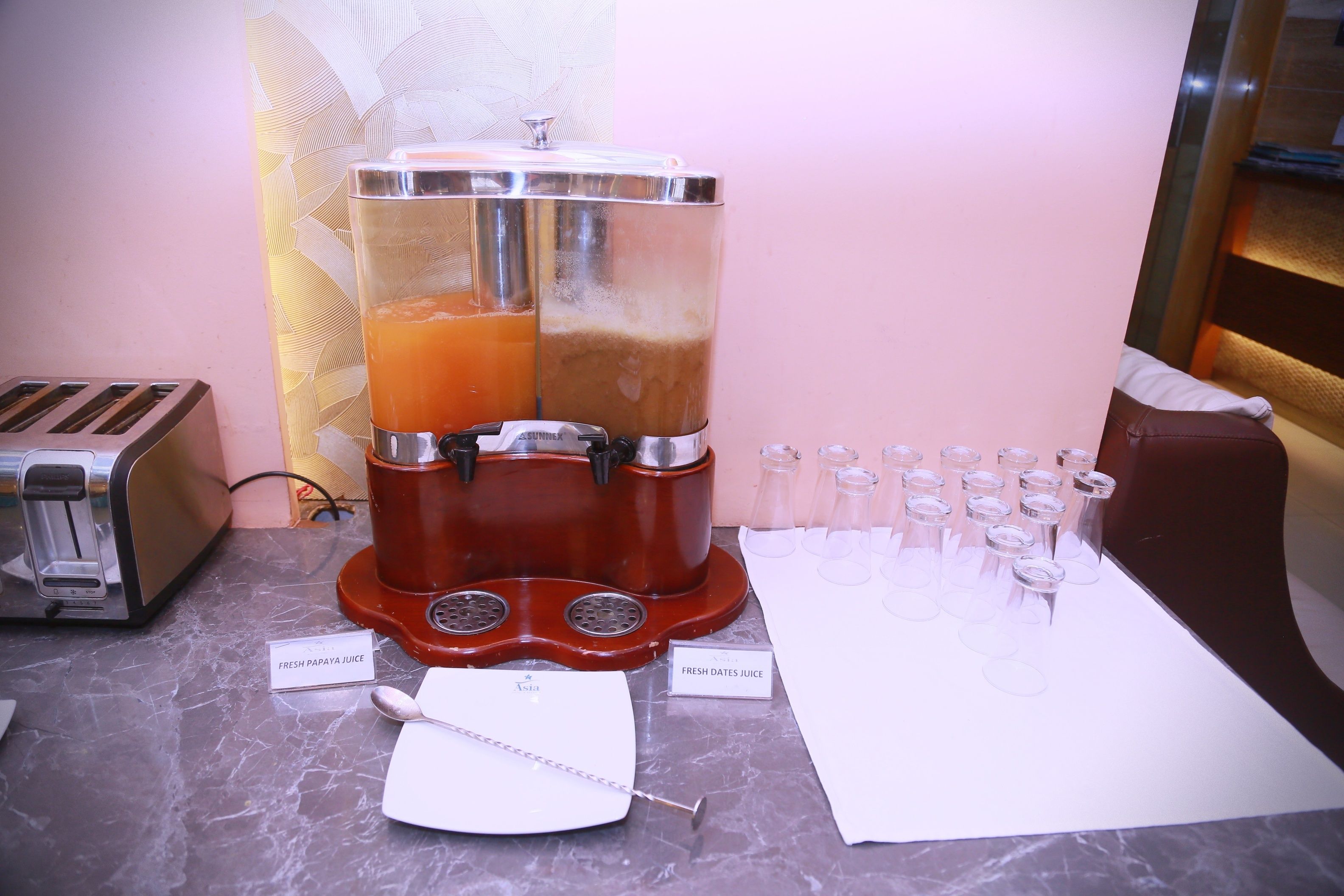 A juice dispenser with two labeled containers, a toaster, and shot glasses lined up on a marble countertop.