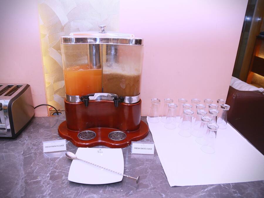 A juice dispenser with two labeled containers, a toaster, and shot glasses lined up on a marble countertop.