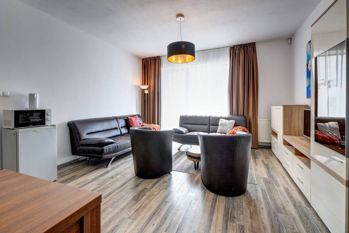 A modern living room with a sectional sofa, two ottomans, a dining table, a kitchenette with a microwave, and a large window with curtains.