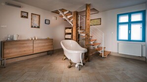 A spacious room with a wooden staircase, a white bathtub with gold legs, a wooden cabinet, and a blue-framed window. - Apartments Ceresnovy Sad (Mengusovce)