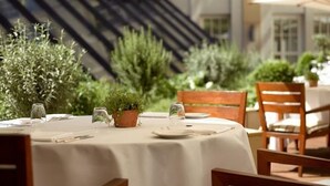 Un espace extérieur aménagé pour prendre les repas, avec une table dressée pour deux personnes, entouré de plantes en pot et avec un escalier en arrière-plan.