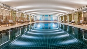 Une grande piscine intérieure avec un sol carrelé, entourée de fauteuils en osier et d’un mur de pierre orné d’une fresque.