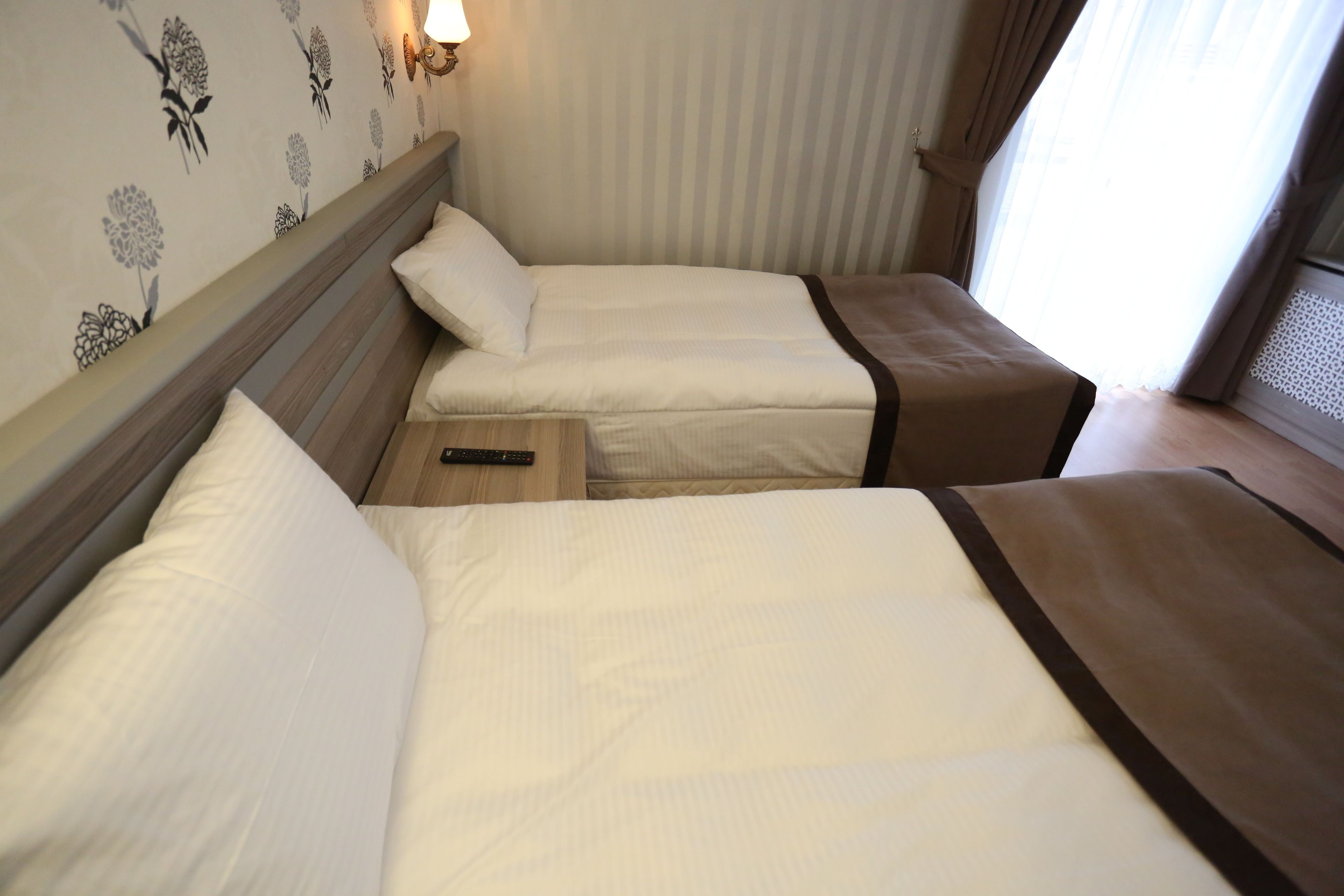 A hotel room with two beds, a nightstand, a lamp, and a window with curtains.