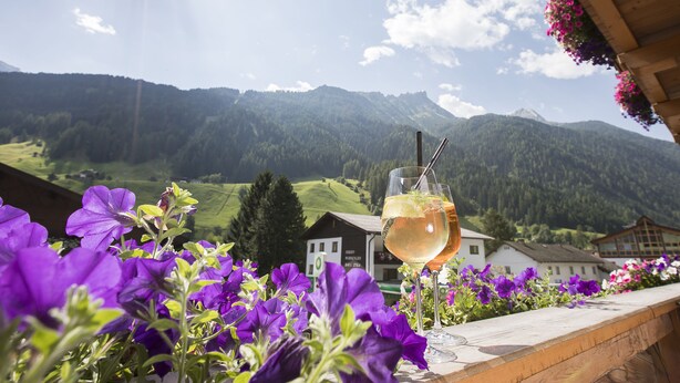 A glass of white wine on a balcony with purple flowers and a mountain view. - Hotel Augarten (Neustift Im Stubaital)