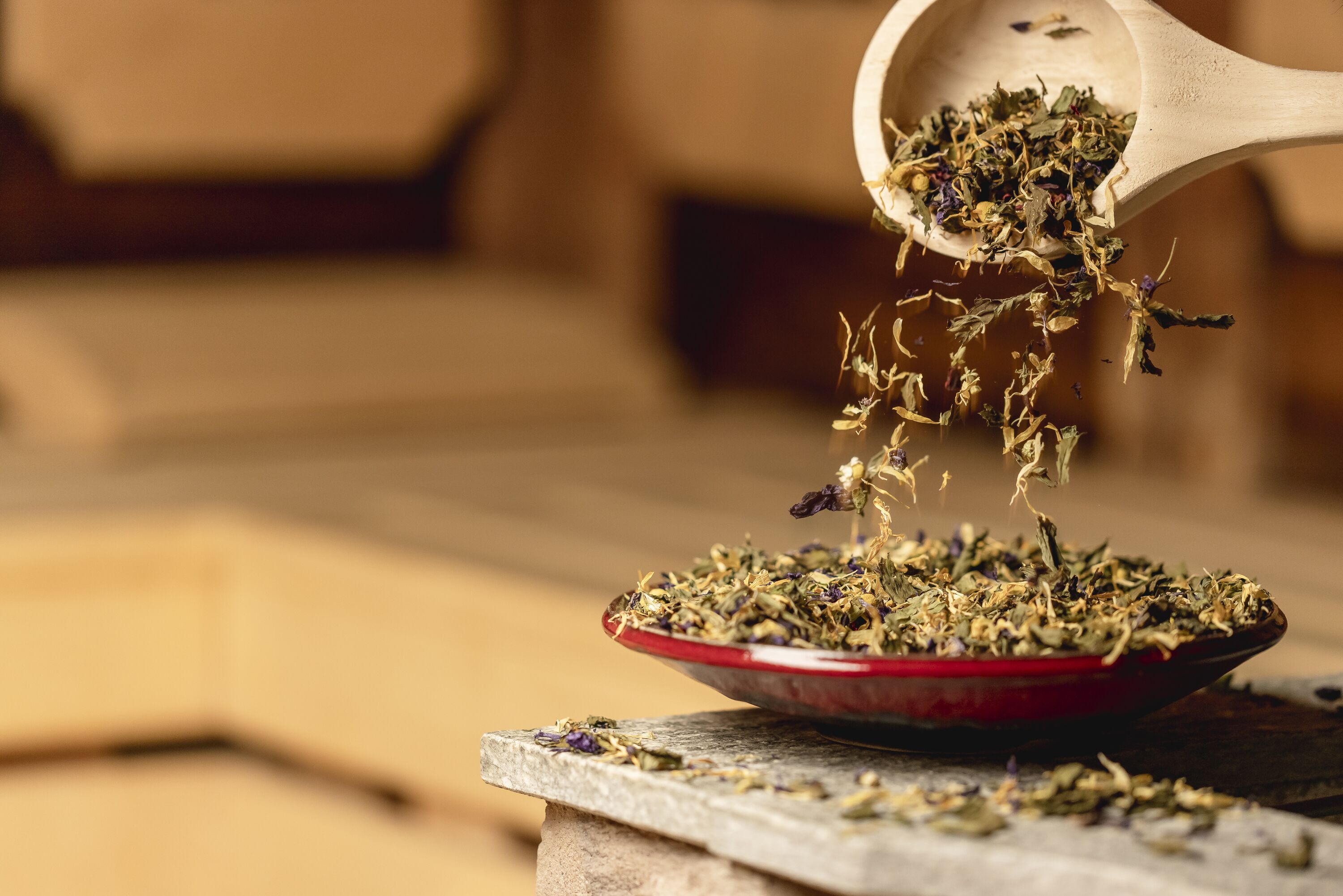 A wooden spoon pouring dried herbs into a red bowl on a stone surface.
