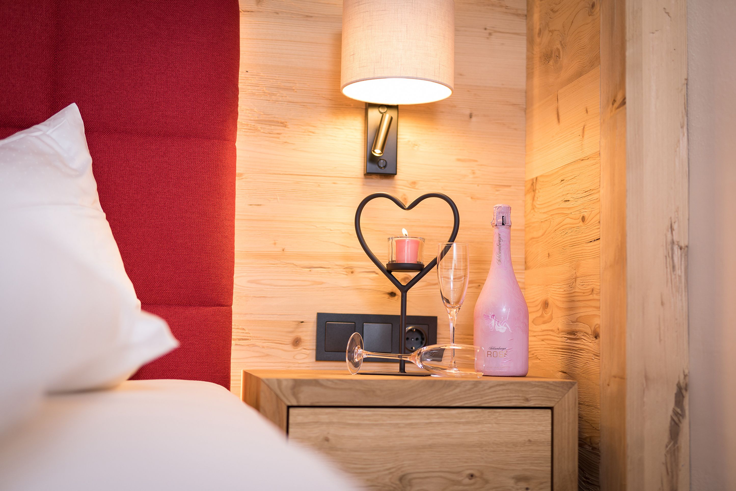 Una mesita de noche de madera bien ordenada, con una lámpara, un candelabro con forma de corazón, una botella de vino y una copa de vino.