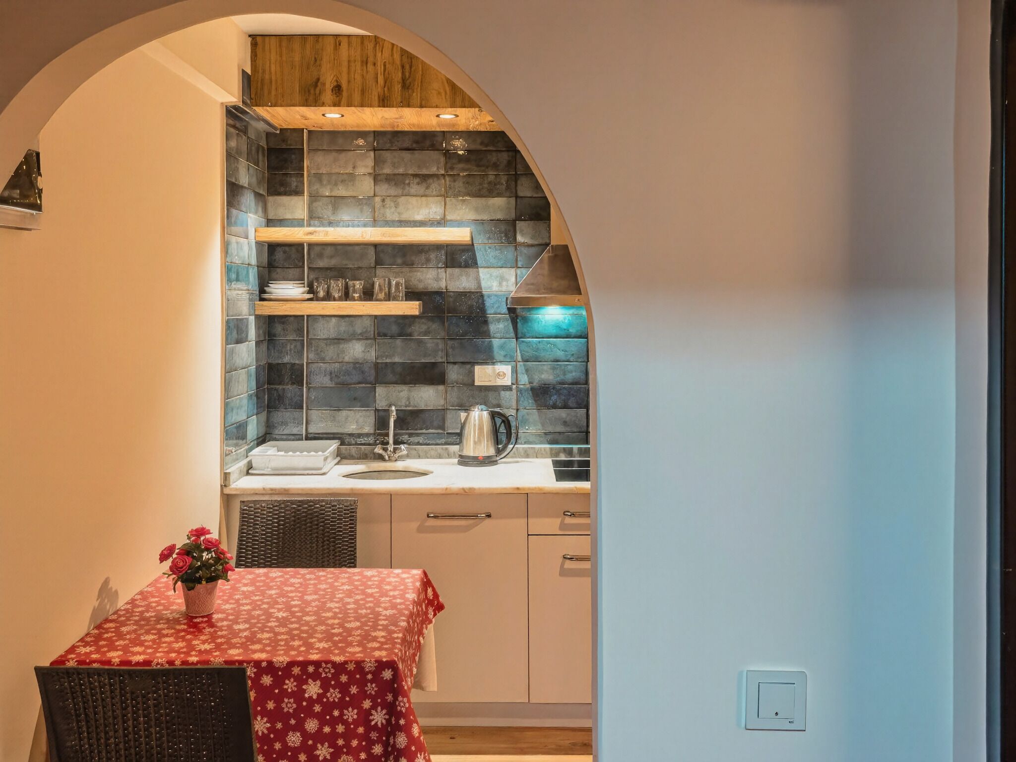 A compact kitchen with a tiled backsplash, a wooden shelf, a sink, and a table with a red tablecloth.