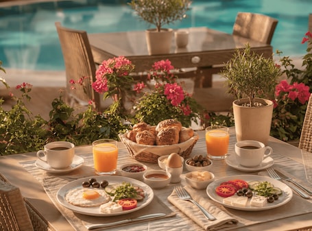 A breakfast setting with a pool view, including croissants, eggs, tomatoes, olives, cheese, and orange juice.. Bella Rose Apart Hotel