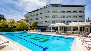 A swimming pool with a clear blue water mark, surrounded by lounge chairs and umbrellas, with a multi-story building in the background. - Hotel Smeraldo (Lignano Sabbiadoro)