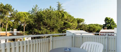 A balcony with a round table and two white chairs, a white fence, and a view of trees and buildings.