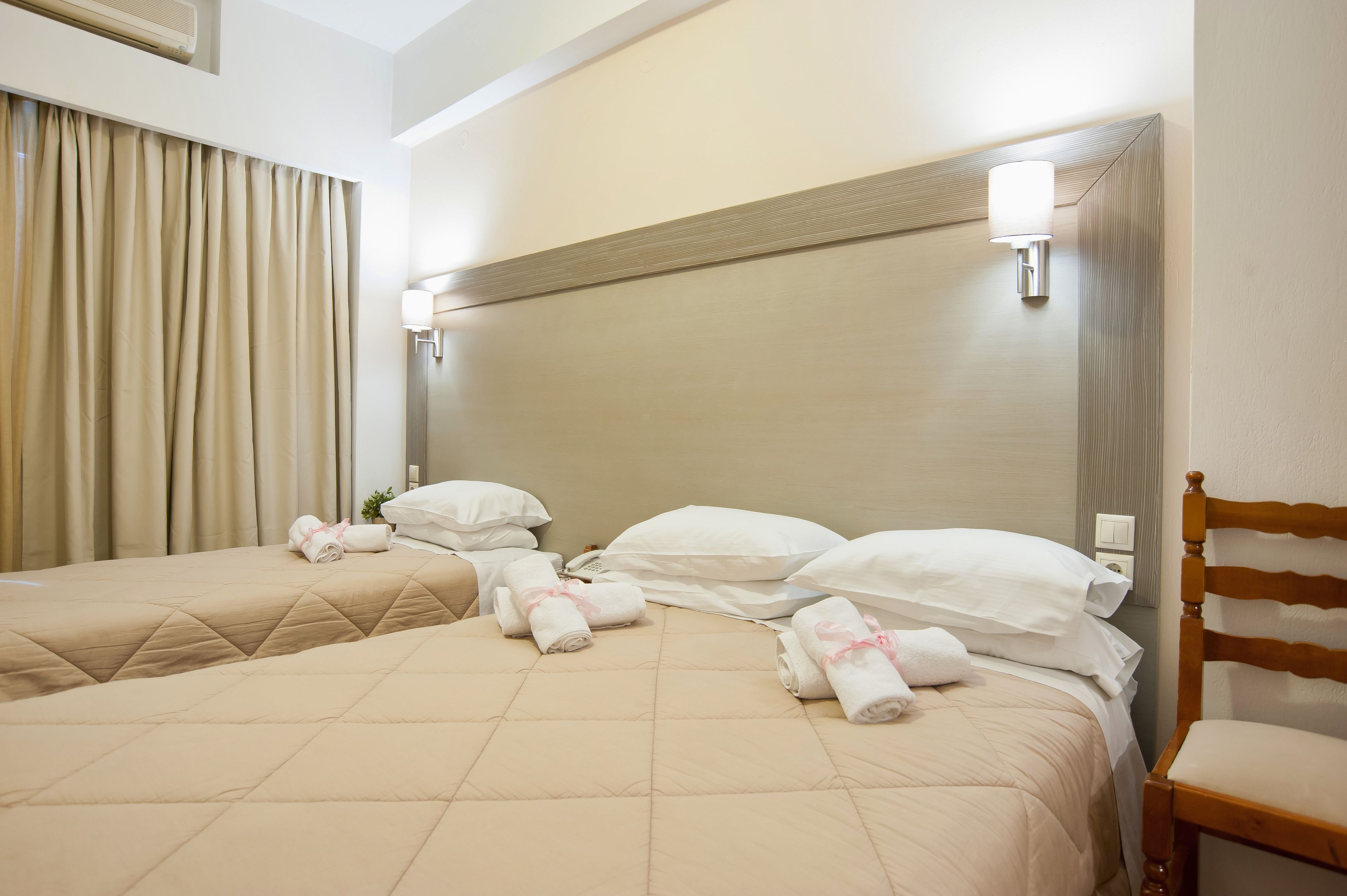 Hotellihuone, jossa on kaksi sänkyä, beigenväriset vuodevaatteet, valkoiset tyynyt ja puinen tuoli.