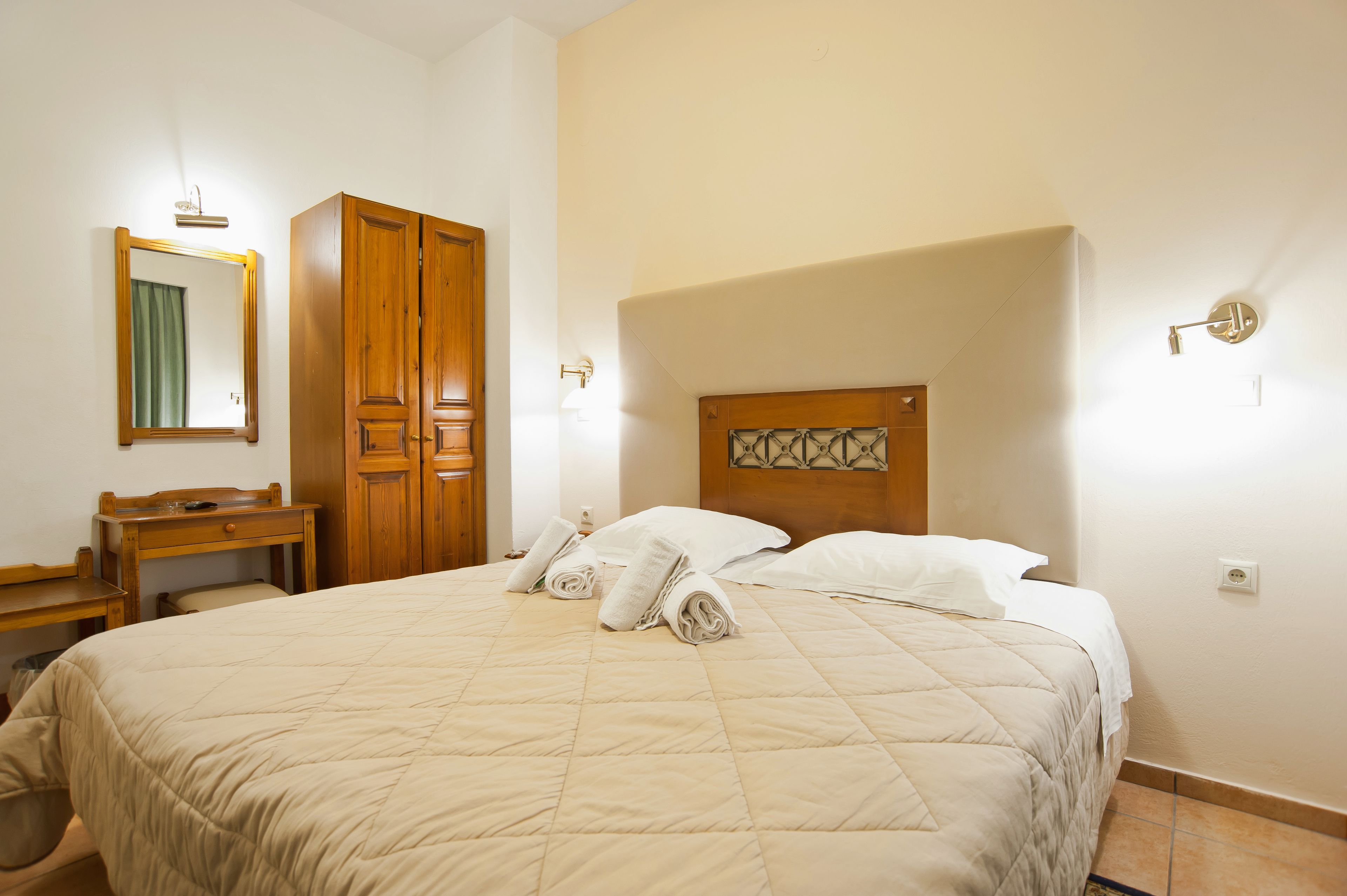 Quarto de hotel com uma cama grande, guarda-roupa de madeira, uma pequena mesa de trabalho e um espelho.