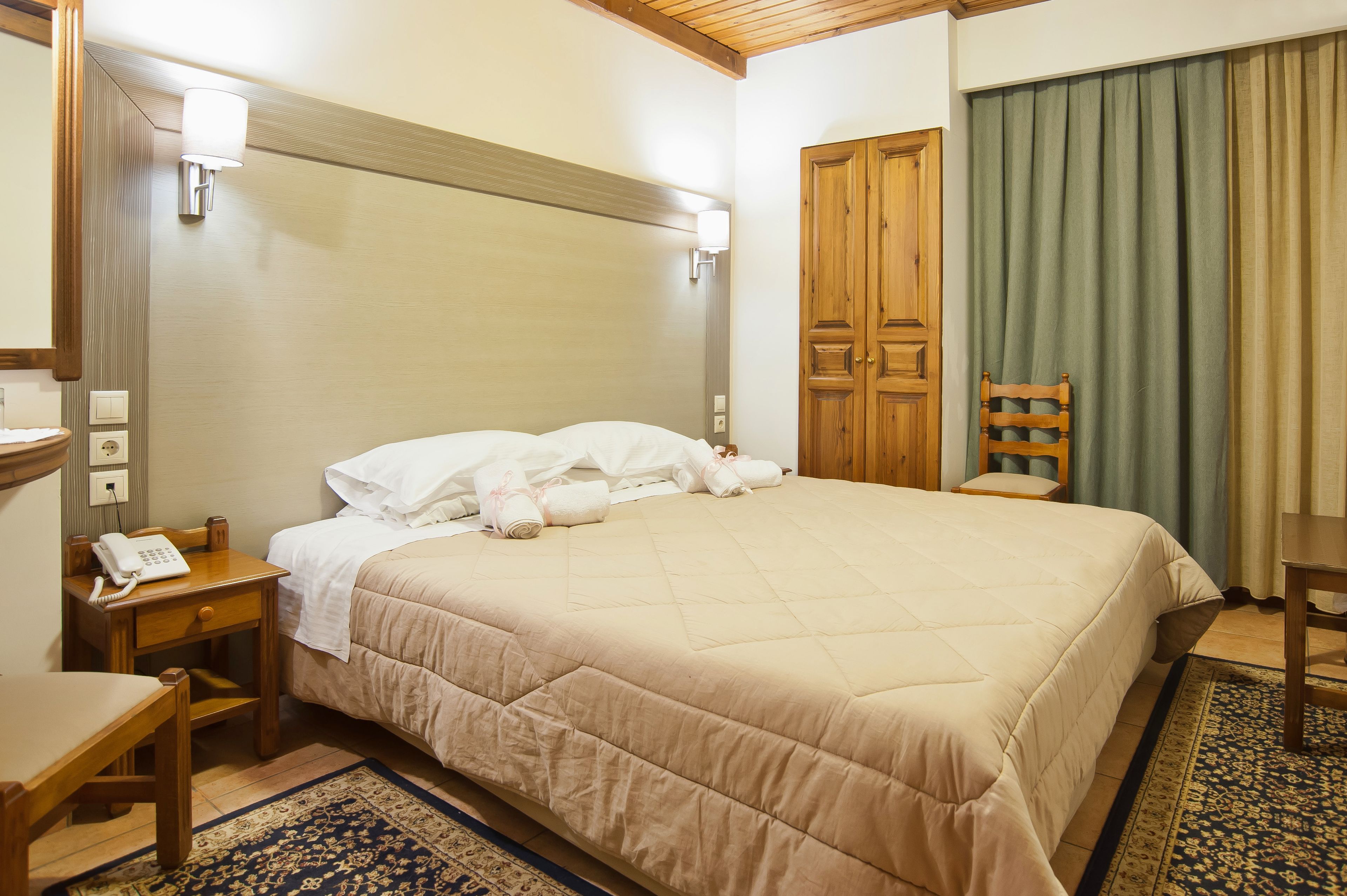 A hotel room with a bed, a wooden nightstand with a telephone, a chair, and a desk.