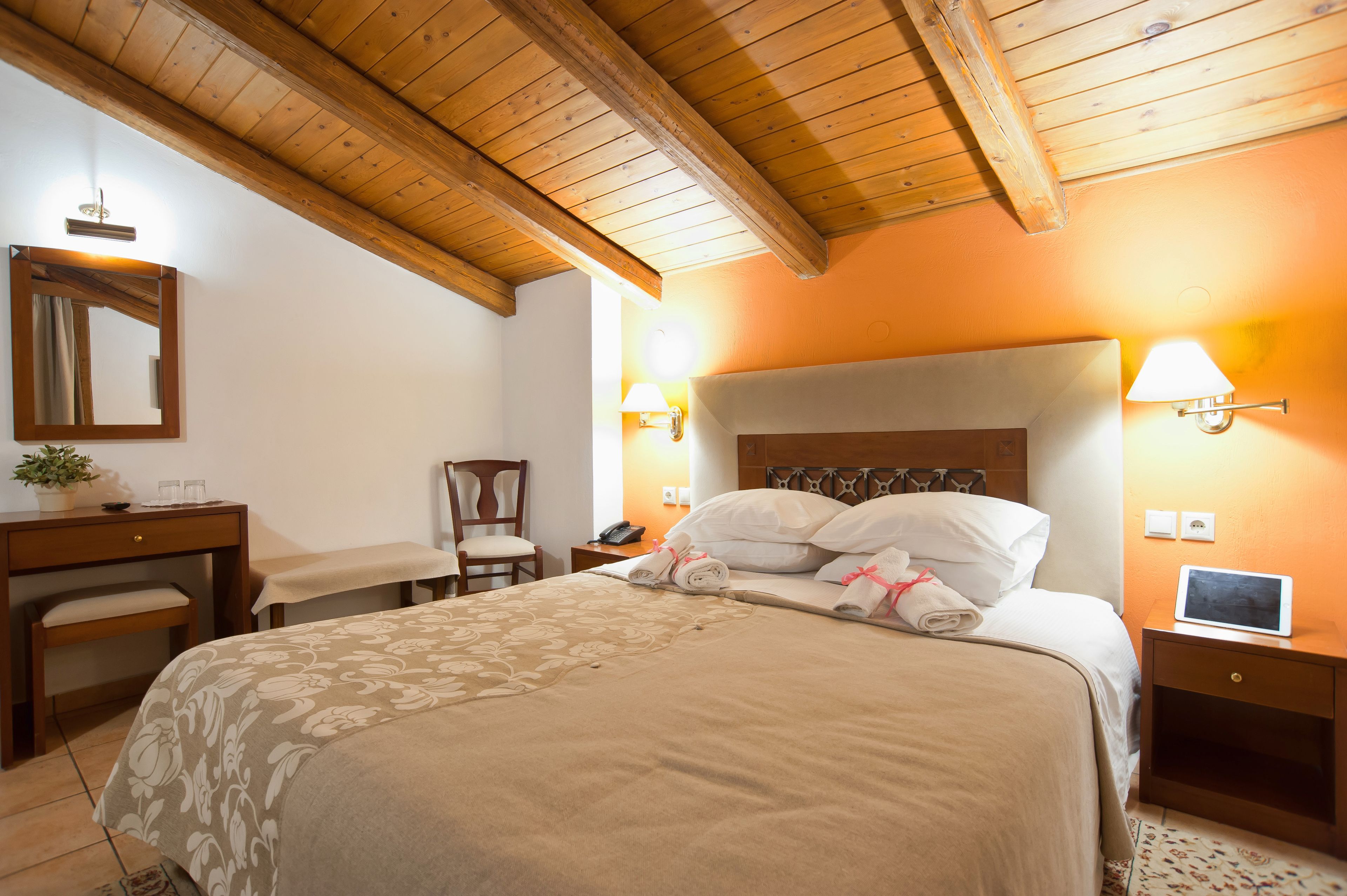 A hotel room with a wooden ceiling, a bed with a floral bedspread, a desk with a chair, and two bedside lamps.