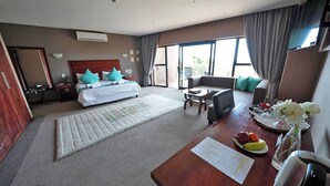 Ein Hotelzimmer mit Bett, einer Couch, einem Schreibtisch mit Obst- und Blumenplatte und Blick ins Freie.