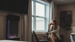 A person sitting by a window in a room with a fireplace, a small table, and a washing machine. - Riverside Suites (Grand Falls - Windsor)