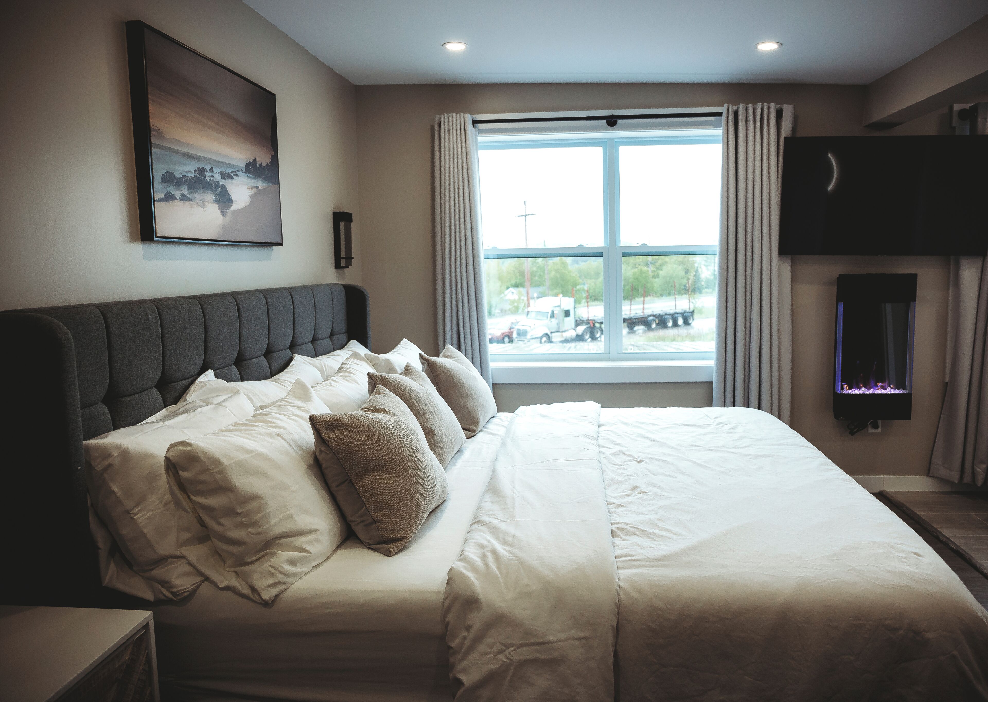 A neatly made bed with white and beige bedding, a large window with curtains, a mounted television, and a painting on the wall.