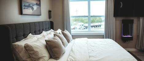 A neatly made bed with white and beige bedding, a large window with curtains, a mounted television, and a painting on the wall.