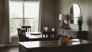 A dining area with a table, chairs, a lamp, a mirror, and a window with curtains. - Riverside Suites (Grand Falls - Windsor)