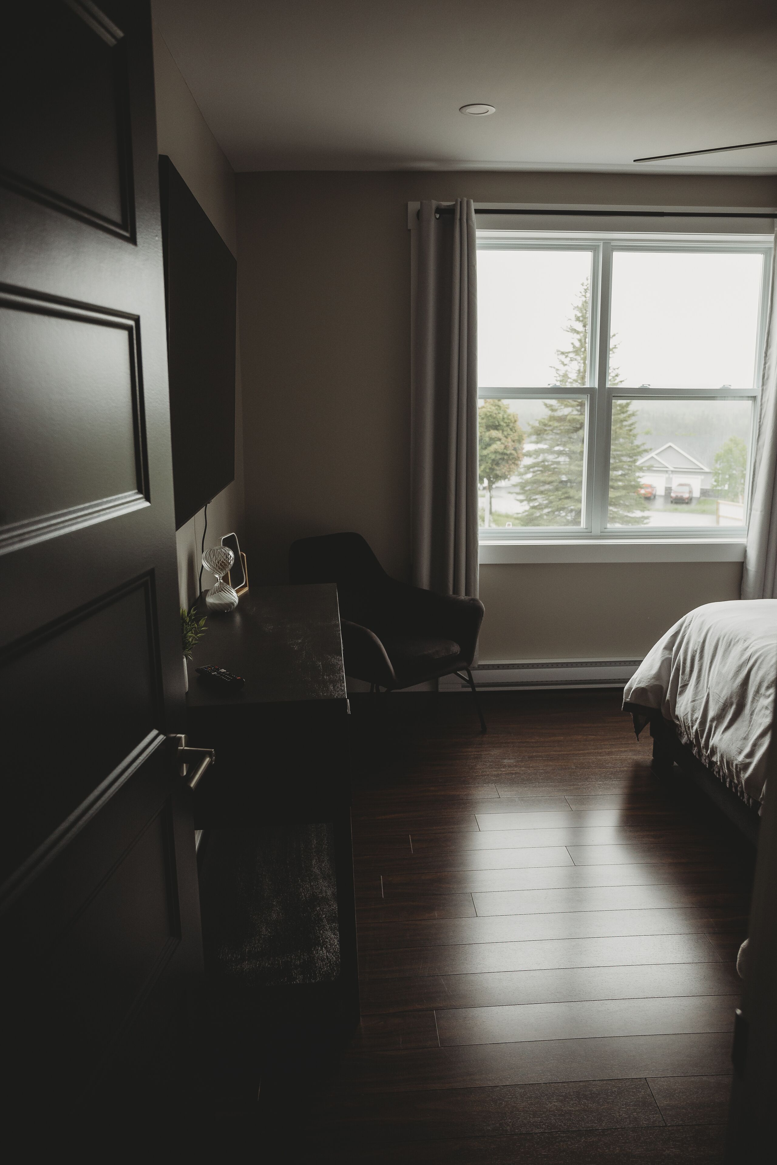 A bedroom with a bed, a desk, and a chair. There is a window with curtains and a view of trees and a house outside.