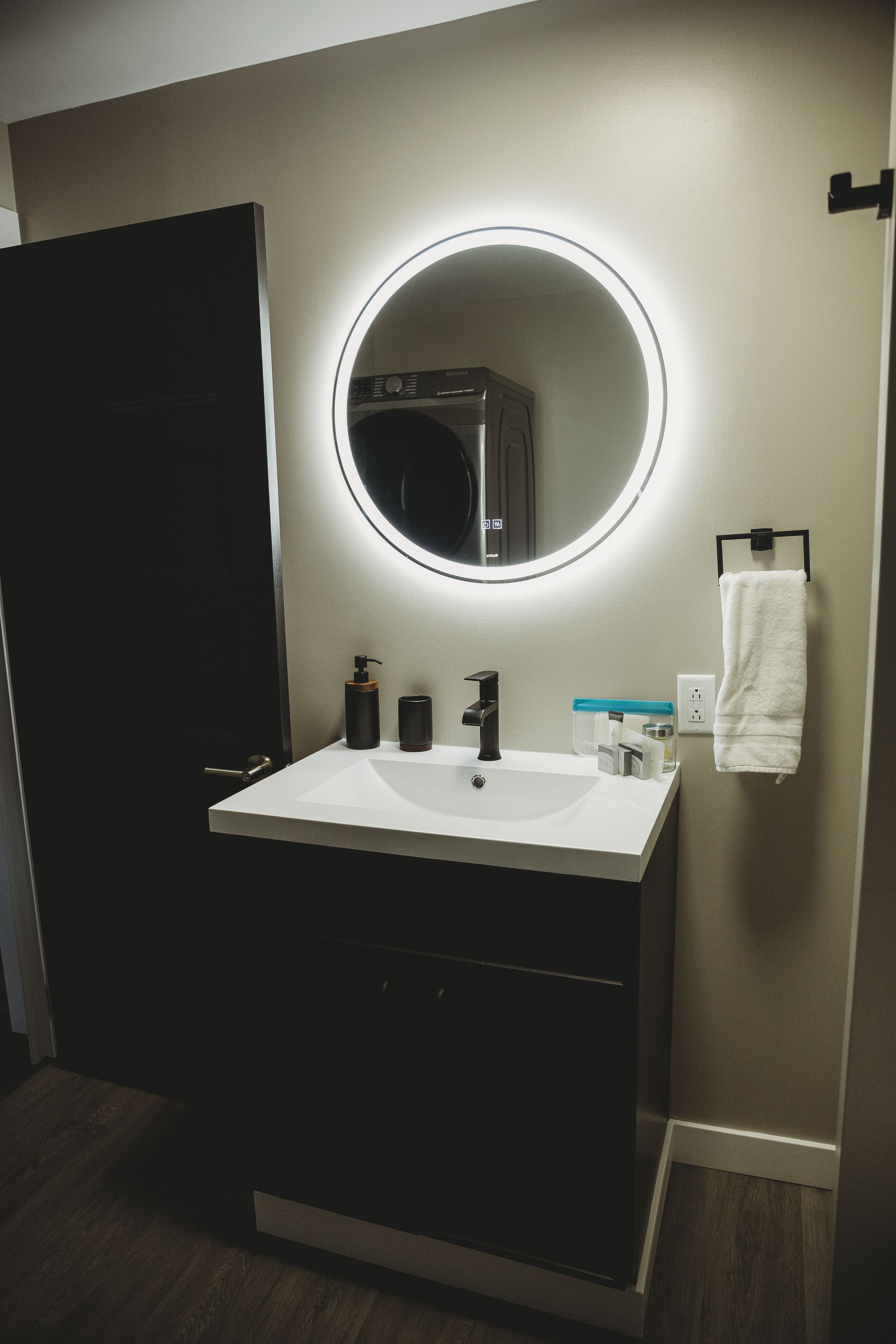 A modern bathroom with a white sink, black faucet, and a round mirror with a light. There are toiletries on the sink and a towel hanging on the wall.