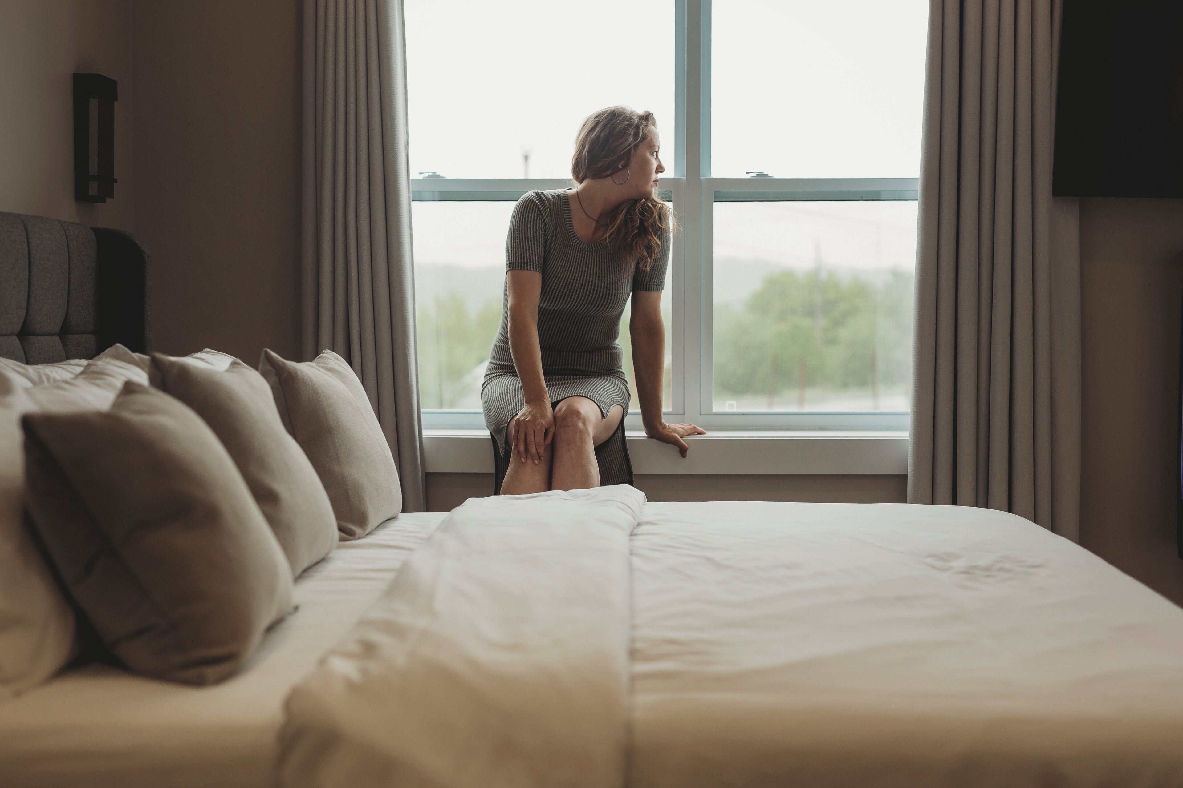 A woman sitting by a window in a bedroom with a bed, pillows, and a wall-mounted TV.