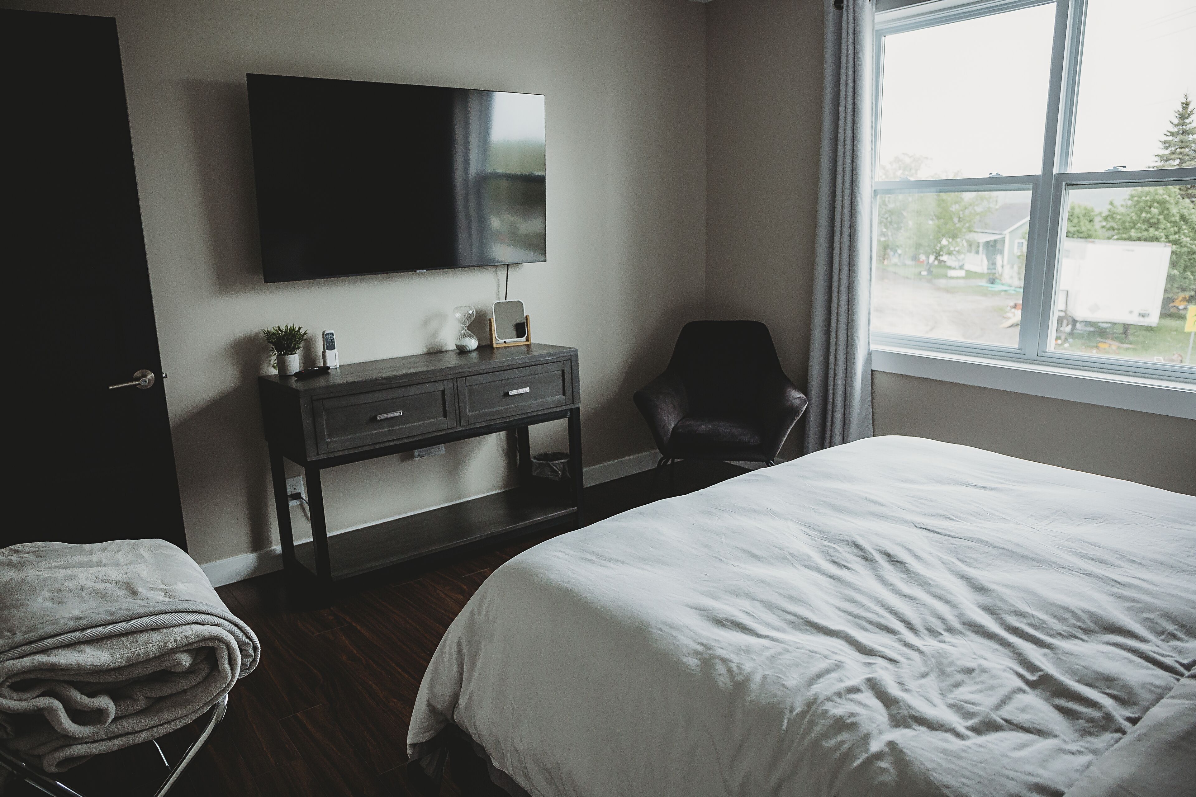 Een slaapkamer met een bed, een aan de muur bevestigde televisie, een stoel en een raam met uitzicht op buiten.