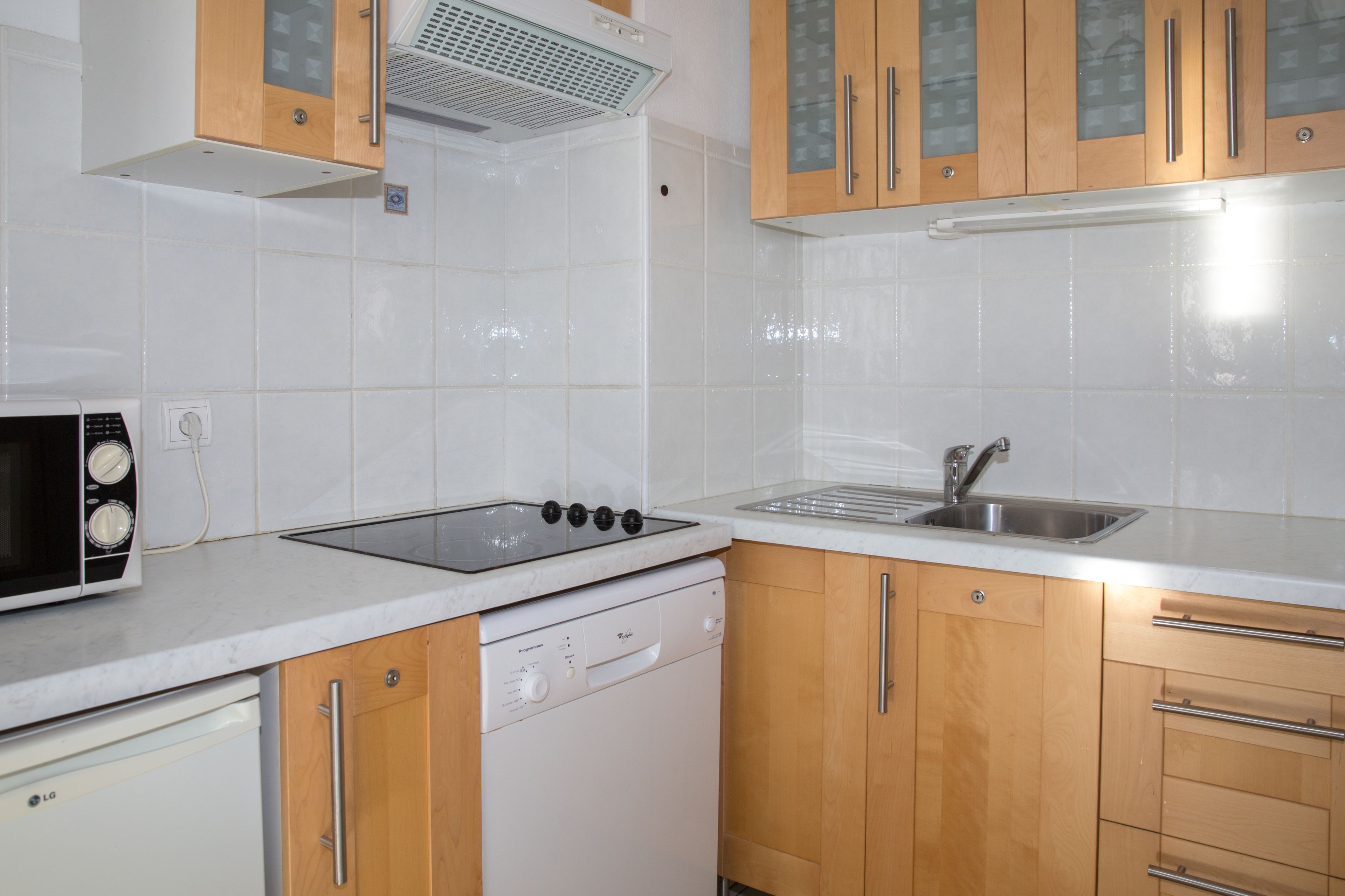Una cocina con azulejos blancos, armarios de madera, microondas, lavavajillas y un fregadero.