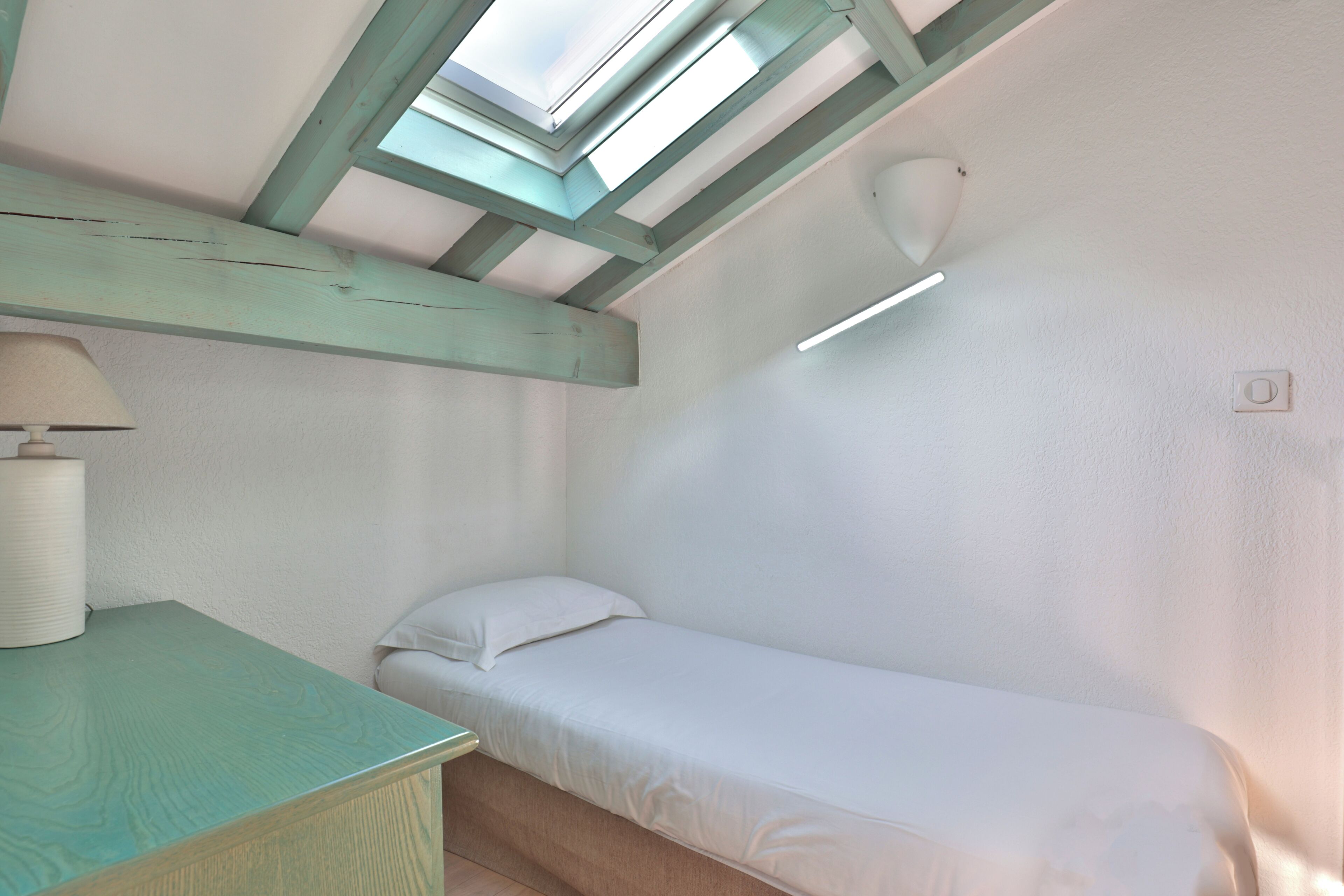 A small, single-bed room with a skylight, a bedside table with a lamp, and a white wall.