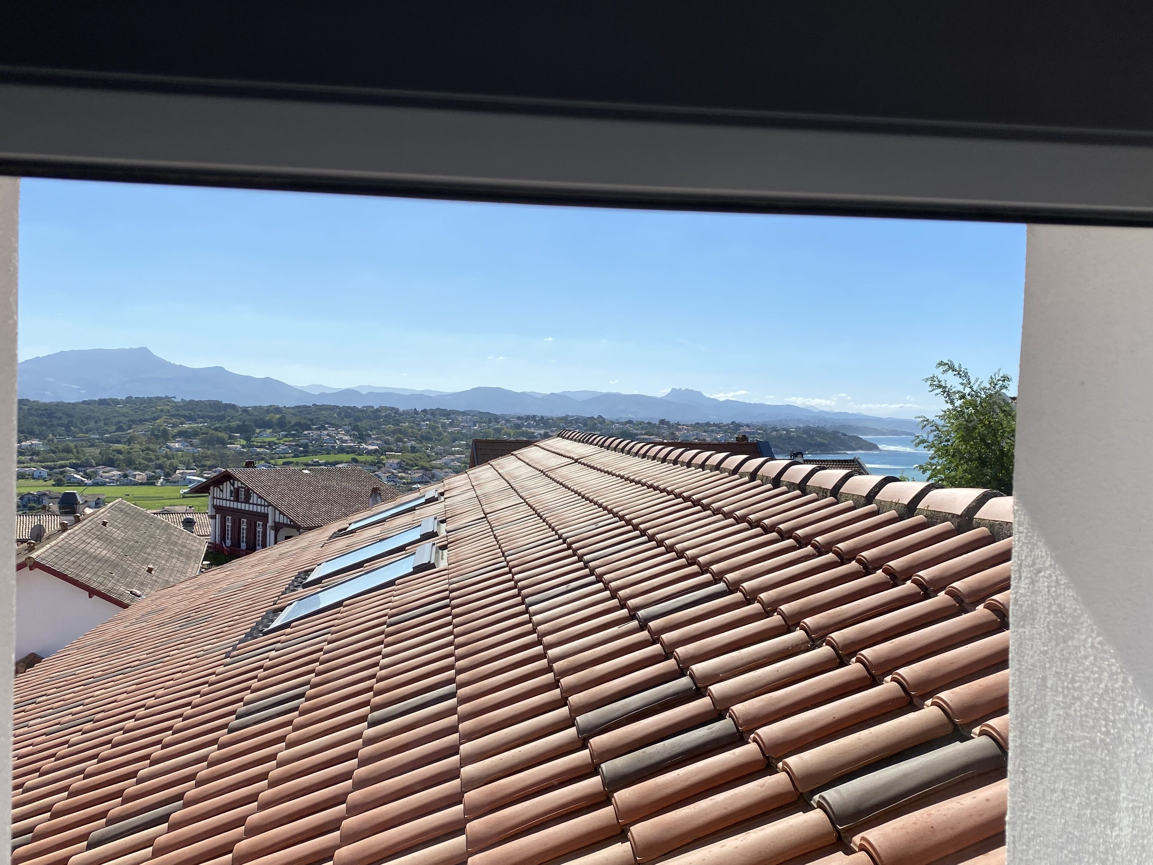 A view from a window overlooking a terracotta-tiled roof, a clear sky, and distant mountains.