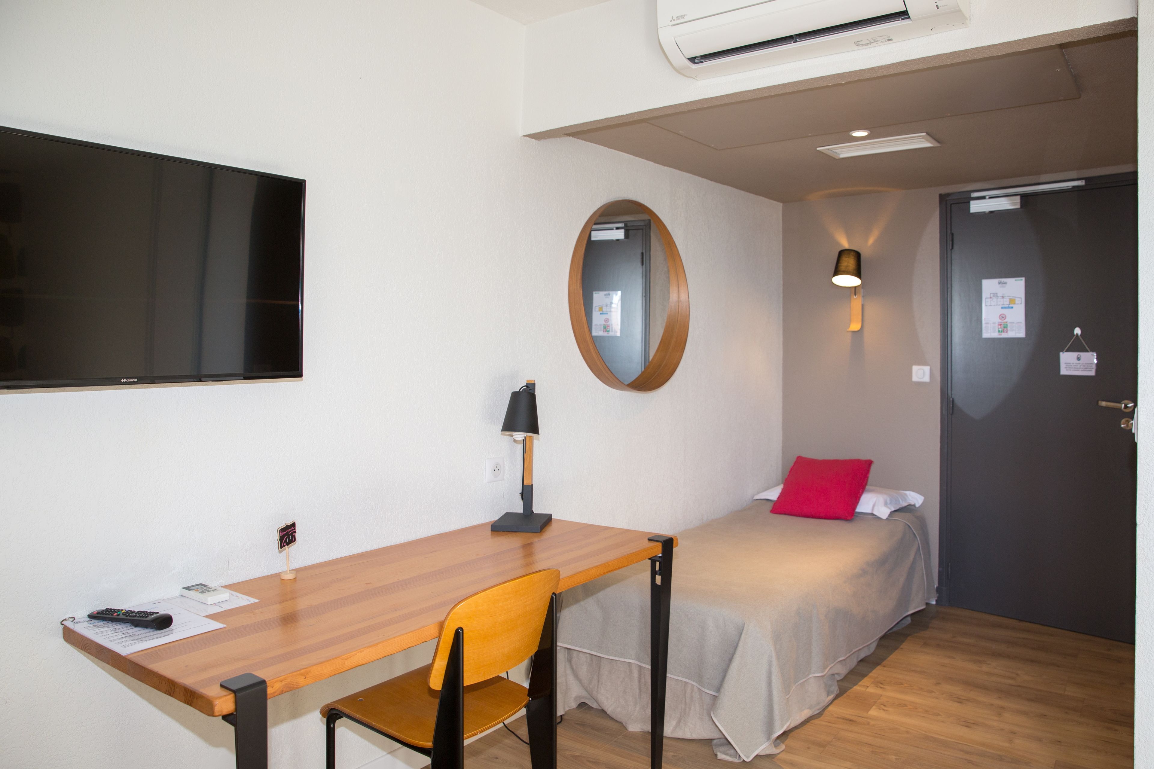 Una habitación con una cama, un escritorio, una silla, un televisor, un espejo y una puerta.