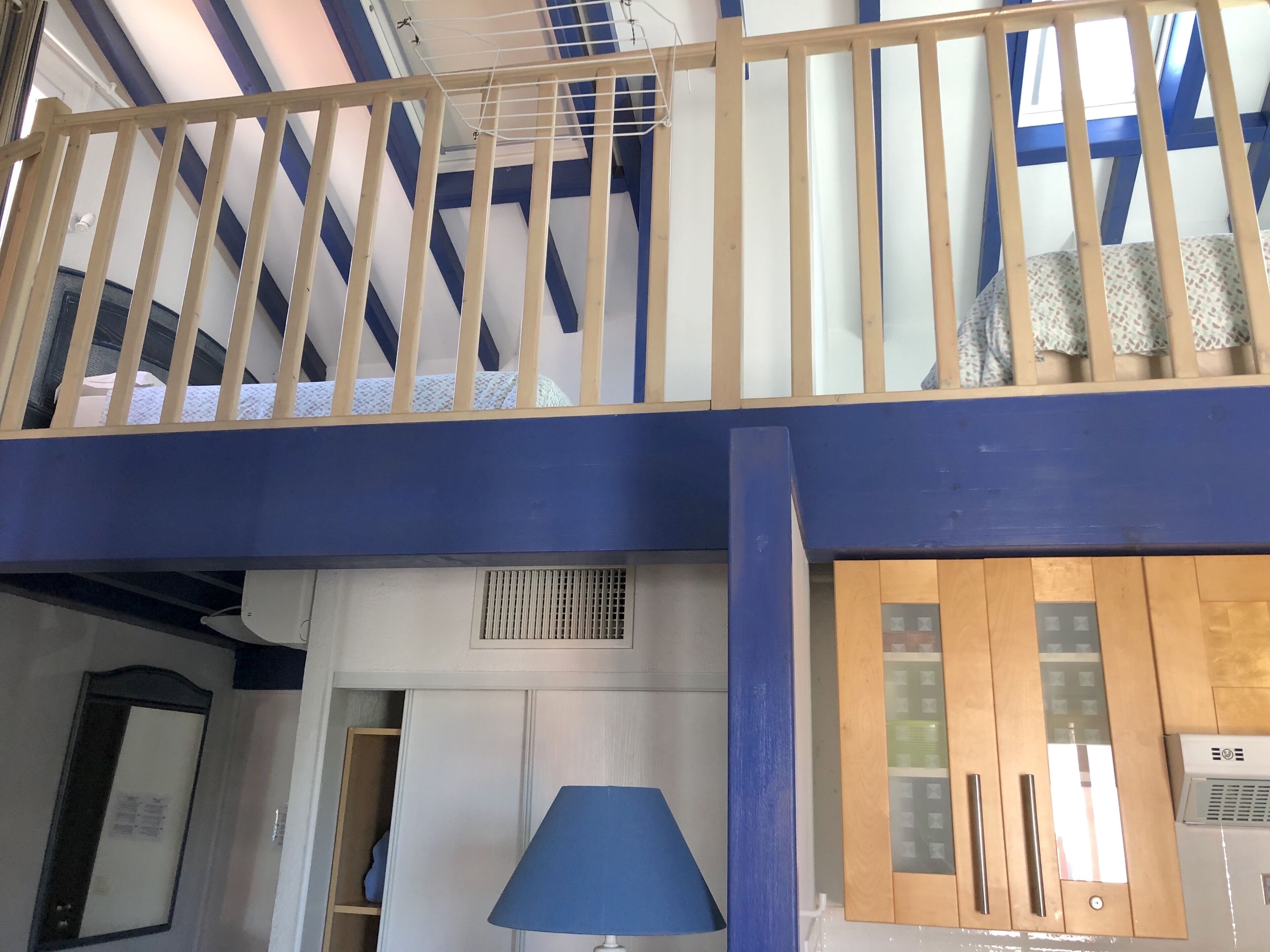 A room with a high ceiling, a wooden balcony railing, a blue lamp, and a kitchen area with wooden cabinets and a refrigerator.