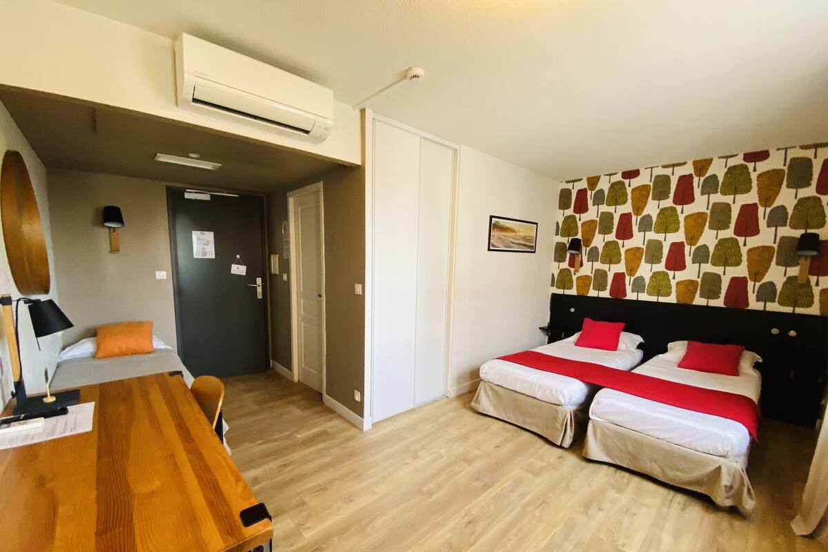 A hotel room with two beds, a wooden desk, an air conditioning unit, and a patterned wall.