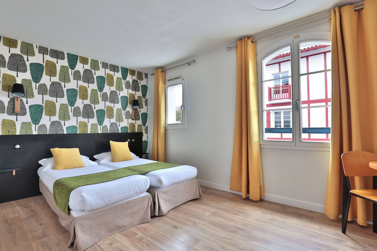 A hotel room with a bed, two bedside tables, a chair, a window with curtains, and a wall with a leaf pattern.