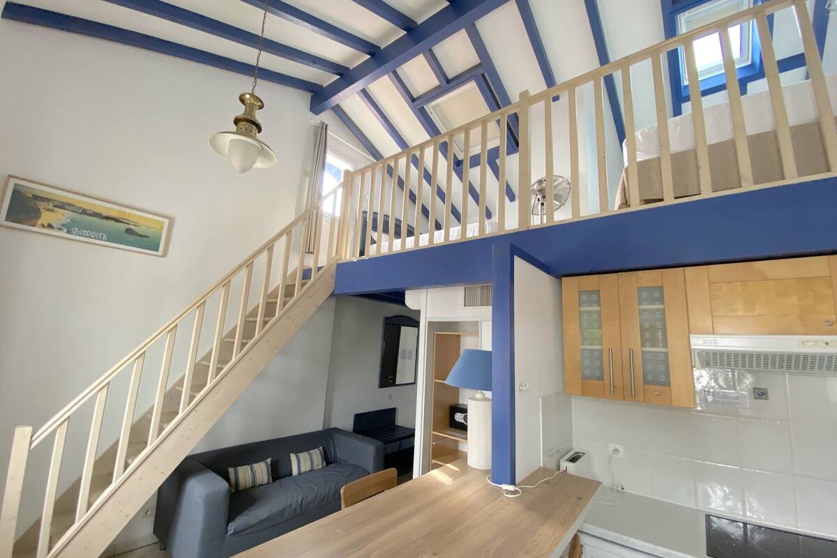 A modern apartment with a wooden staircase, a dining area, a kitchen with a washing machine, and a view of the sea through a window.