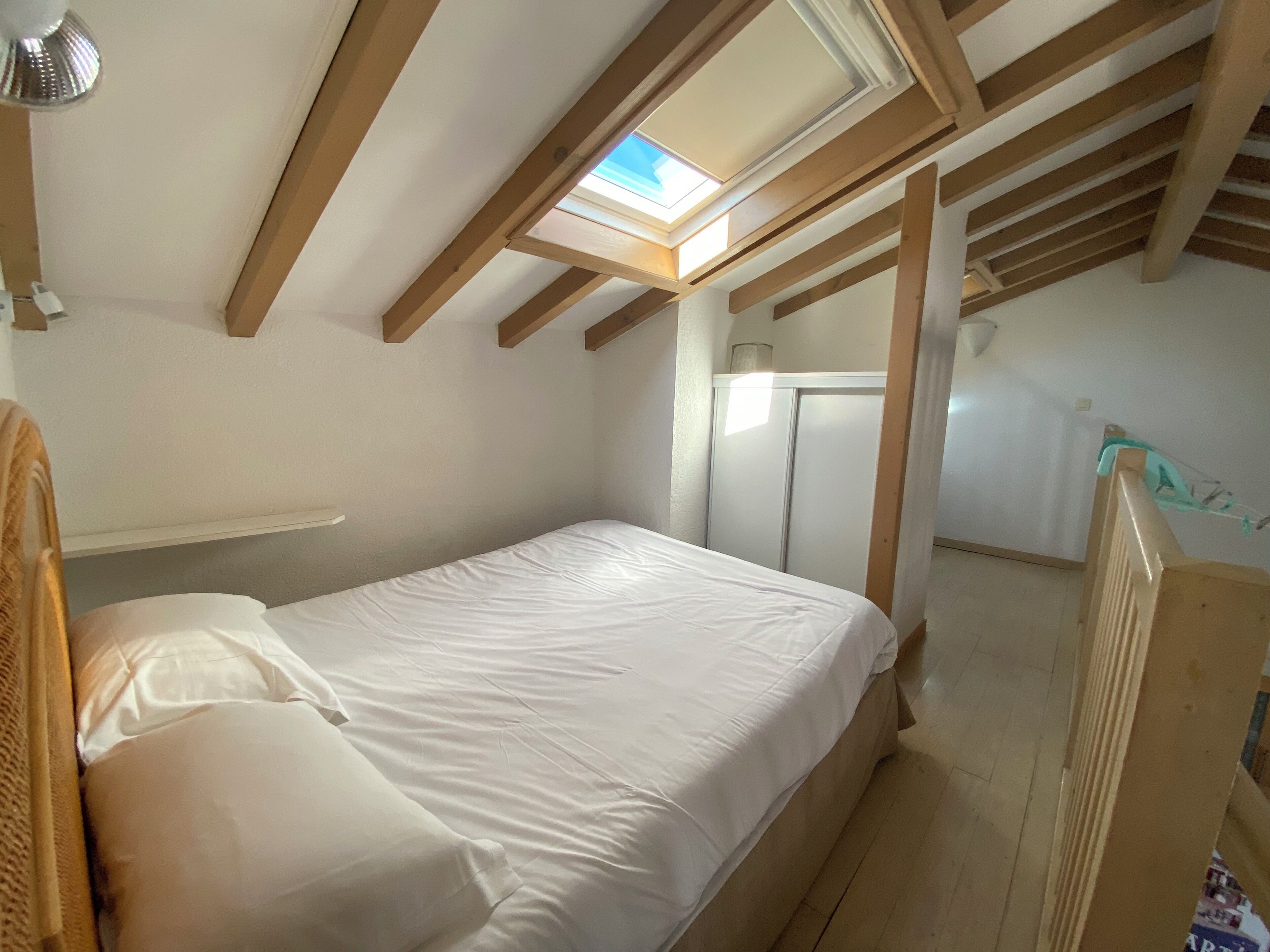 A single bed in a room with a sloped ceiling and skylight, wooden beams, a wardrobe, and a small shelf.