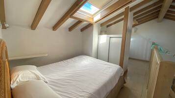 A single bed in a room with a sloped ceiling and skylight, wooden beams, a wardrobe, and a small shelf.
