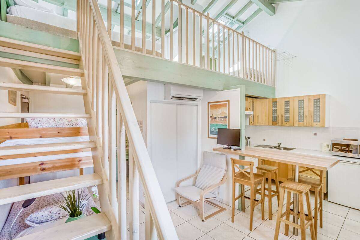 A modern kitchen with a wooden staircase, a dining area with stools, and a view of the outdoors.
