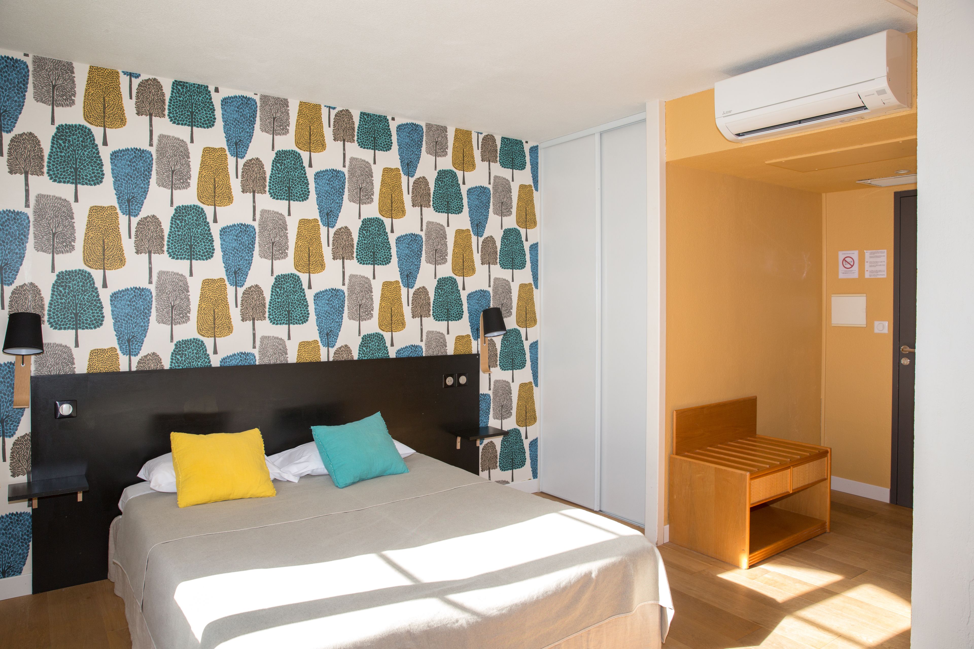 Une chambre à coucher comprenant un lit, une table de chevet, un climatiseur fixé au mur et un petit banc en bois.
