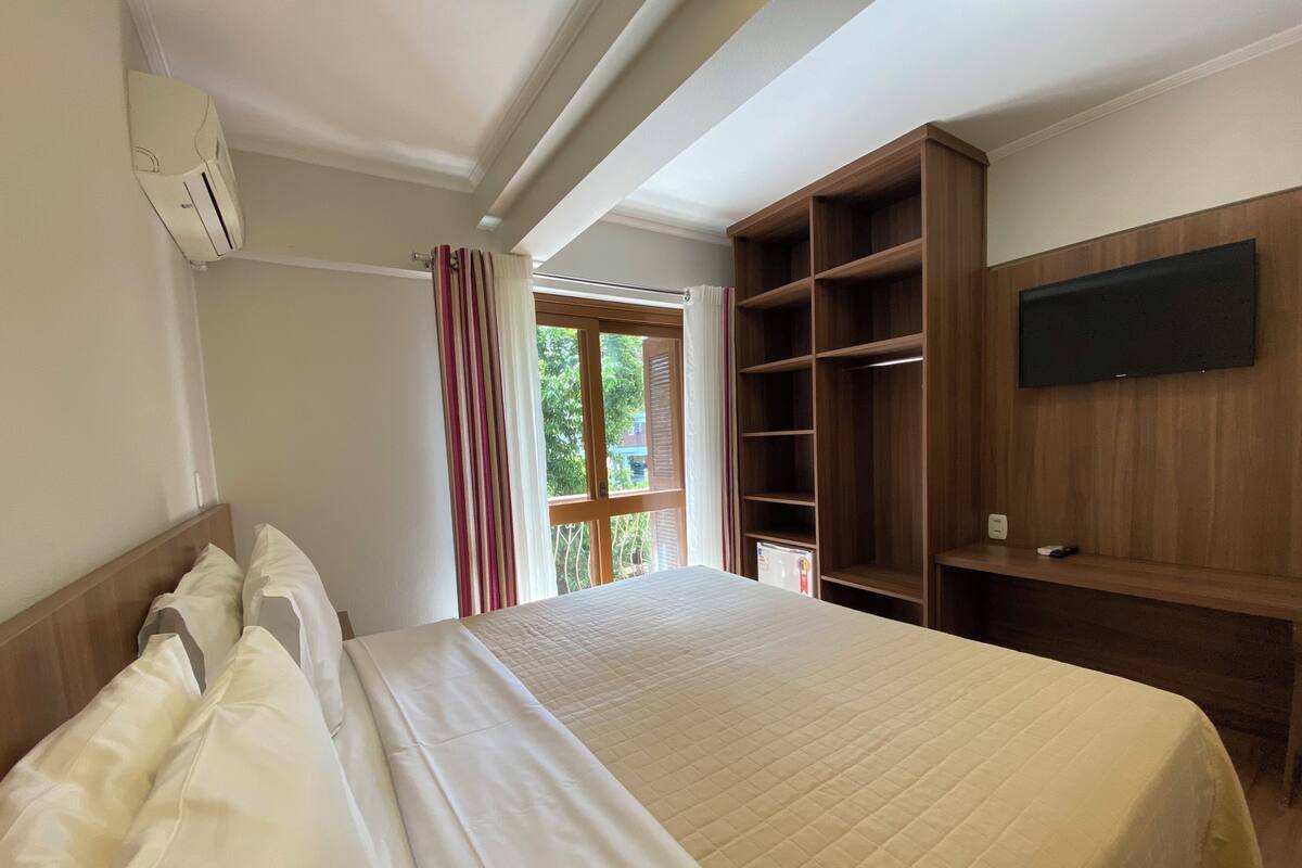 A hotel room with a bed, a TV mounted on the wall, a wooden wardrobe, and a window with curtains.