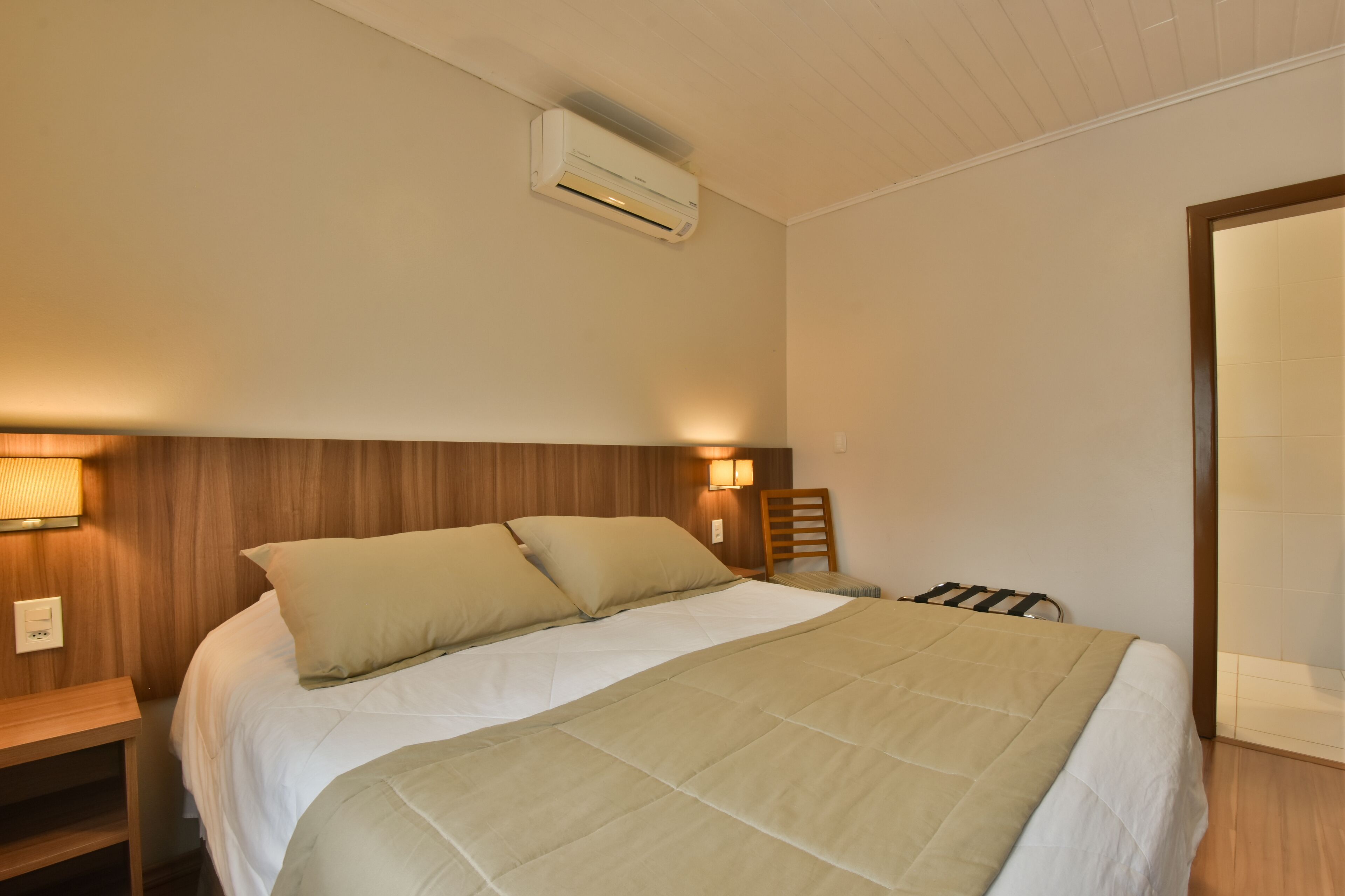 A neatly made bed with a beige quilt, a wooden headboard, and a wall-mounted air conditioner.
