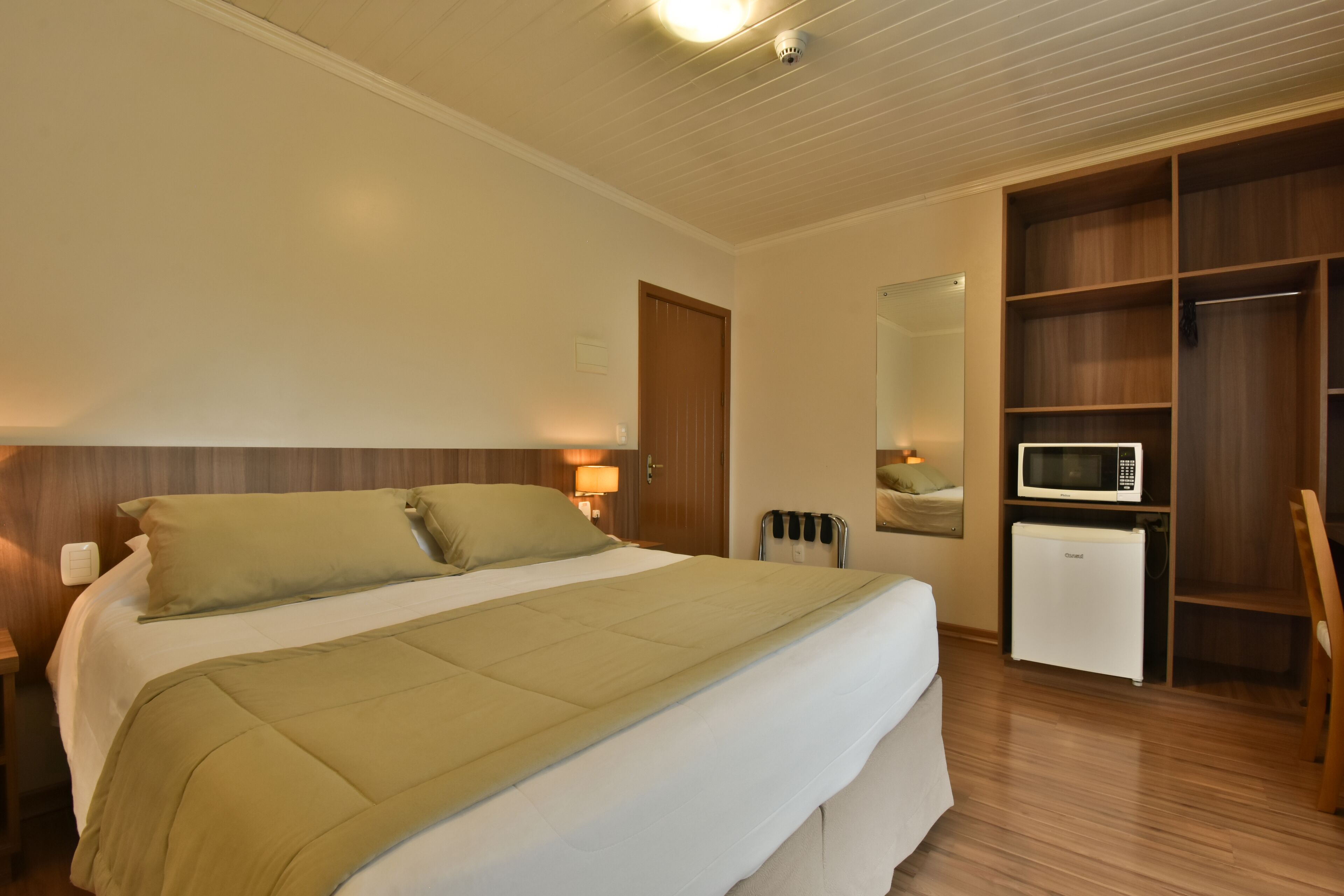 A hotel room with a bed, a wooden wardrobe, a small refrigerator, and a mirror.