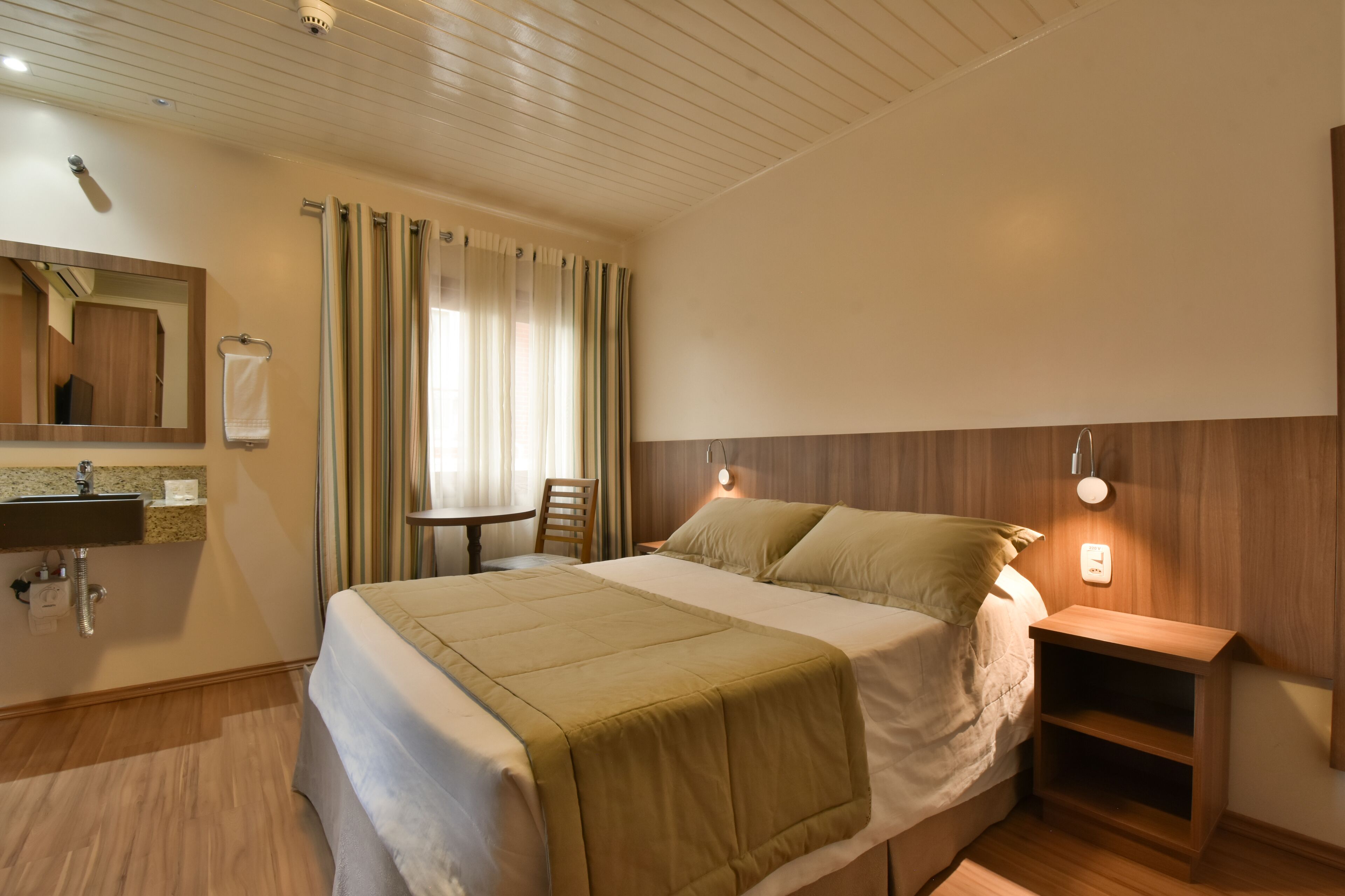 A hotel room with a bed, a bedside table, a nightstand, a mirror, a sink, and a window with curtains.