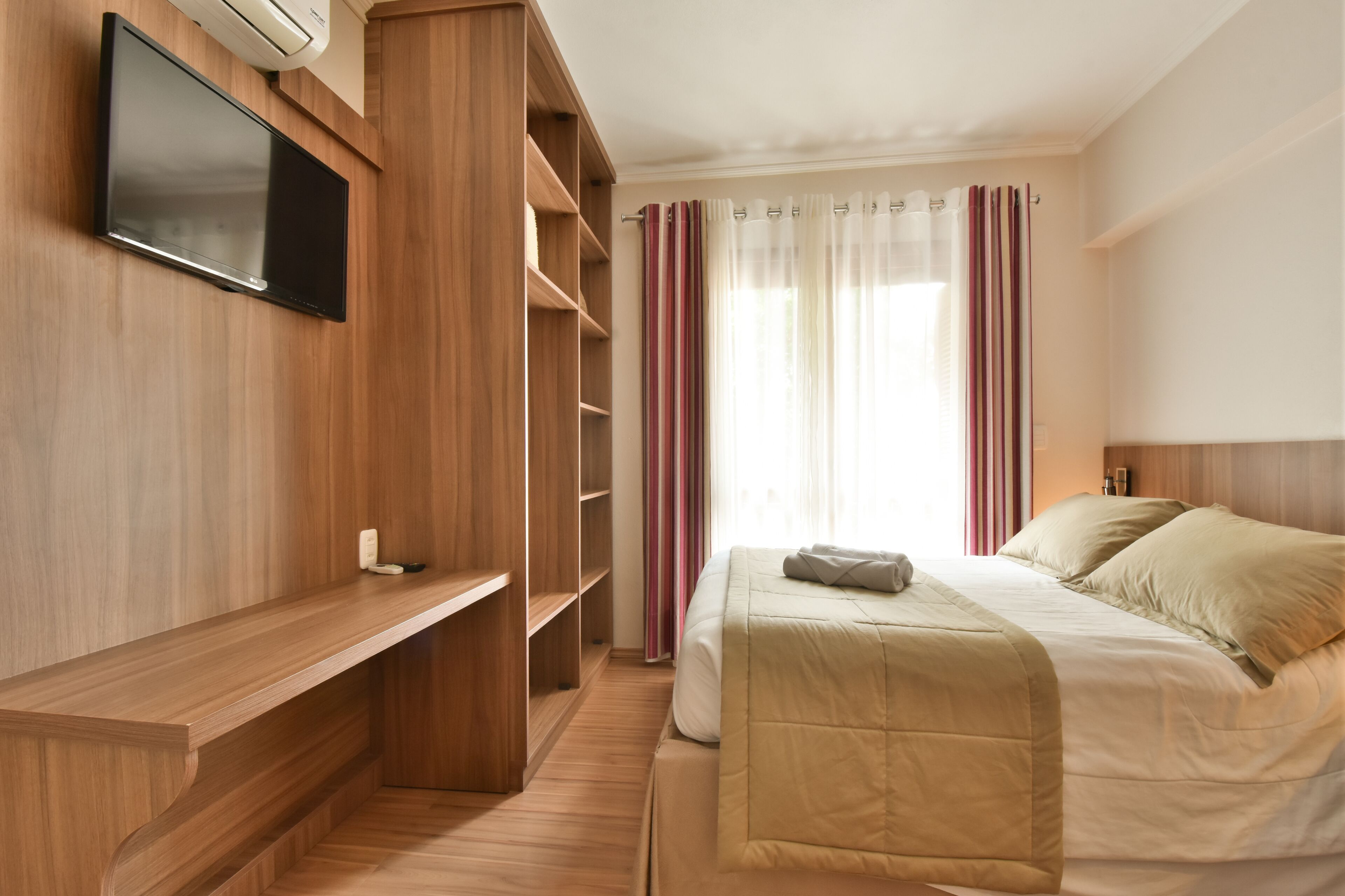 Habitación de hotel con un armario de madera, un televisor de pantalla plana montado en la pared, una cama con ropa de cama color beige y una ventana con cortinas traslúcidas.