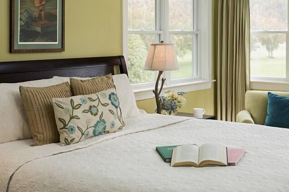 A neatly made bed with a white comforter and several pillows, a bedside table with a lamp and a cup, and a window with curtains.