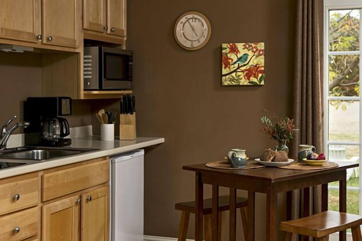 A kitchen with wooden cabinets, a microwave, and a sink. There's a clock on the wall and a painting of birds. A table with a vase and a plate of fruit is in front of a window.