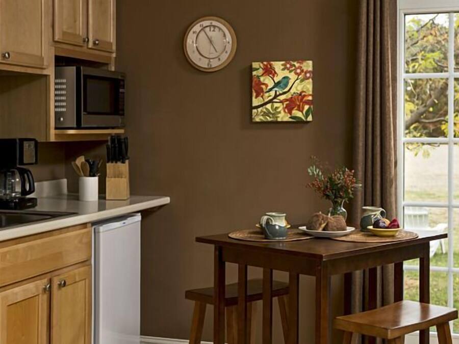 Une cuisine avec des armoires en bois, un four à micro-ondes et un évier. Il y a une horloge au mur et un tableau représentant des oiseaux. Une table avec un vase et une assiette de fruits est placée devant une fenêtre.