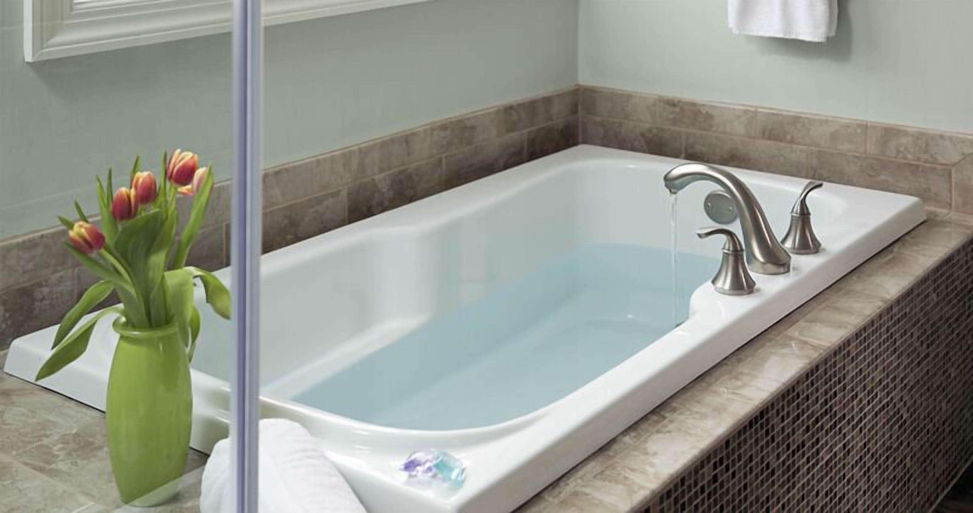 A bathroom with a rectangular bathtub, a green vase with flowers, and a towel hanging on the wall.
