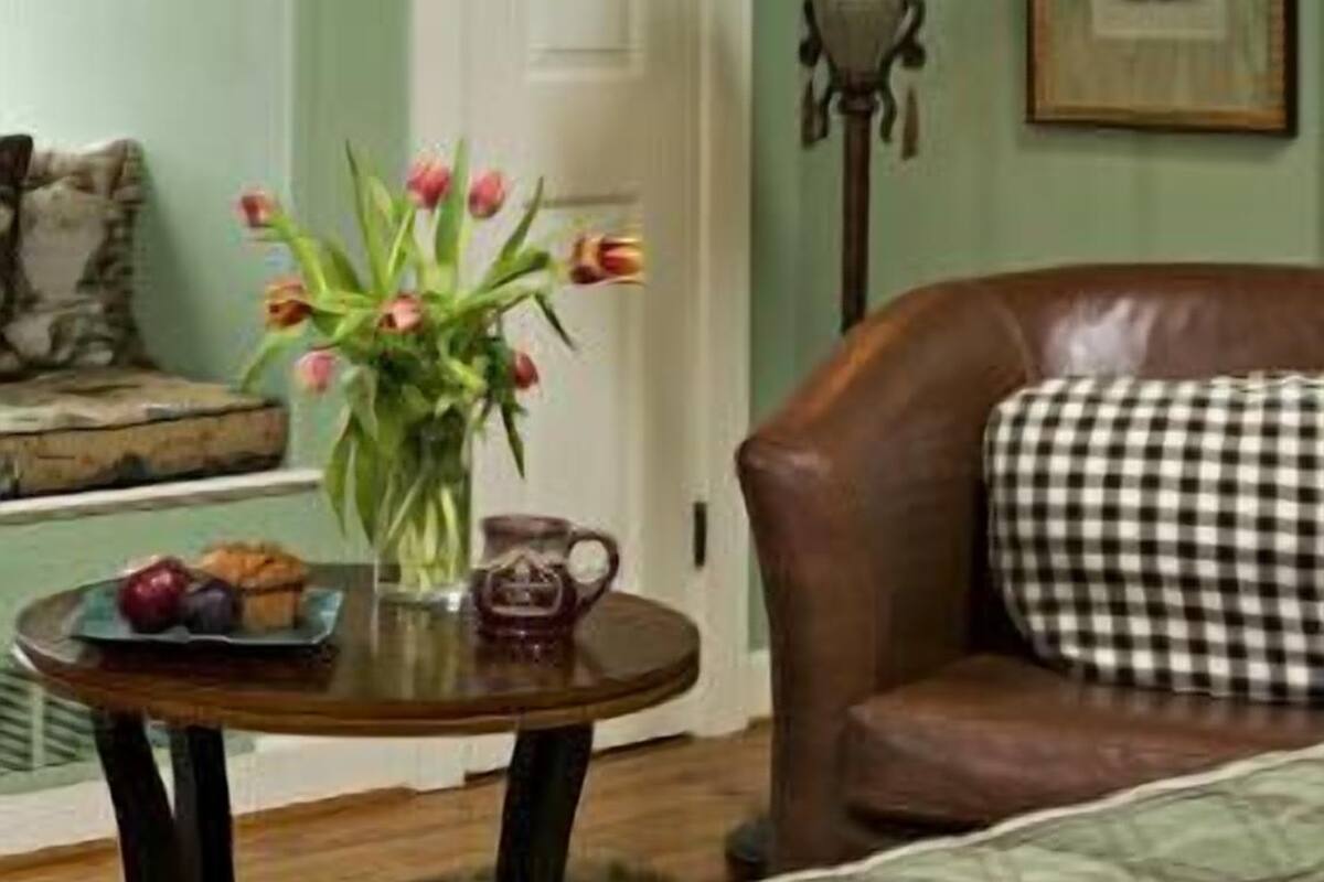 A cozy living room with a brown leather sofa, a small round table with a vase of flowers, a lamp, and a framed picture on the wall.