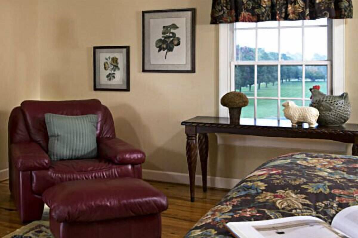 A room with a leather armchair, a floral-patterned bed, a table with decorative items, and framed artwork on the wall.